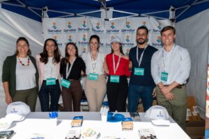Siete personas sonrientes frente a un stand de Servatur, con cordones y vestimenta informal, y con artículos de marca sobre la mesa.