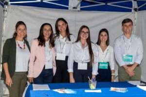 Seis personas están de pie sonriendo detrás de una mesa azul bajo una carpa, llevando etiquetas con sus nombres.