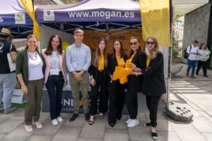 Un grupo de personas vestidas con ropa profesional se sitúa delante de un stand con el nombre "mogan.es", algunos de ellos con americanas negras y camisas amarillas, posando para una foto al aire libre.