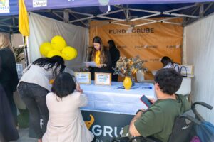 Puesto con la marca "Fund Grube", globos, jarrón amarillo y gente interactuando. Una mujer está detrás de la mesa leyendo un documento.