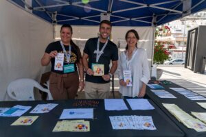 Tres personas en un stand bajo una carpa, sonriendo y sosteniendo panfletos. En la mesa de enfrente hay folletos, bolígrafos y volantes.
