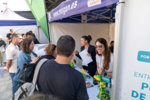 Personas interactuando en un stand al aire libre con folletos y artículos promocionales. Una mujer con gafas de sol sonríe a los visitantes.