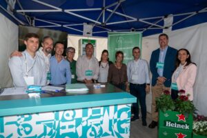 Un grupo de personas juntas bajo una carpa azul en un evento, con pancartas y una mesa que muestra la marca Heineken.