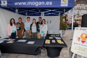 Seis personas posando bajo una carpa azul junto a una mesa con folletos y un cartel de ADL Mogán.