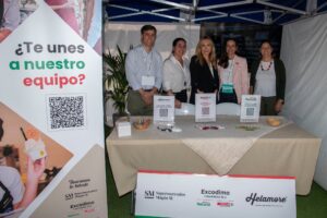 Un grupo de cinco personas se encuentra detrás de una mesa en un stand promocional con materiales informativos y un cartel que invita a las personas a unirse a su equipo.