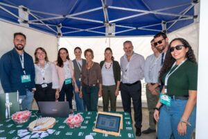 Un grupo de personas se encuentra bajo una carpa con dosel azul en un evento, con una computadora portátil y materiales informativos en una mesa frente a ellos.