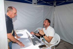 Dos hombres conversaban en una mesa con una computadora portátil, dentro de una estructura similar a una tienda de campaña.