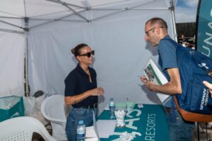 Una persona con gafas de sol está de pie en una mesa bajo una carpa y habla con otra persona que sostiene una fotografía. Hay sillas y material promocional a su alrededor.