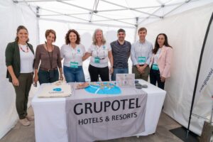 Siete personas de pie dentro de una carpa blanca en una mesa promocional de Grupotel Hotels & Resorts, con bolsas de mano y folletos exhibidos.
