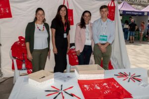 Cuatro personas de pie en un stand con bolsas rojas y bolígrafos sobre una mesa, asistiendo a un evento al aire libre.
