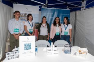 Cinco personas de pie en un stand sonriendo a la cámara, con carteles de "Seaside Collection" y materiales promocionales exhibidos.