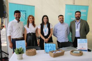 Cinco personas de pie juntas detrás de una mesa con cestas y material promocional en una carpa. Detrás de ellas se muestran dos pancartas verticales.
