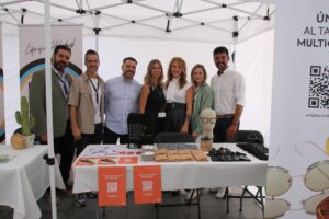 Siete personas están juntas bajo un dosel blanco en un stand con varios artículos sobre la mesa, incluidos folletos, vasos y un cactus.