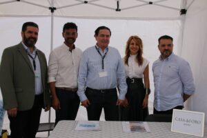 Cinco personas paradas juntas bajo un dosel, vestidas con ropa informal de negocios, con una mesa rotulada "CASA del ORO joyería" en primer plano.