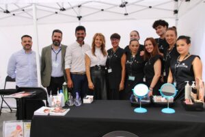 Un grupo de once personas, de pie y sentadas bajo un dosel blanco, posan detrás de una mesa que muestra productos de belleza y espejos.