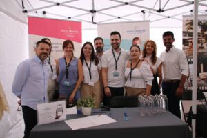 Un grupo de nueve personas se encuentra de pie detrás de una mesa bajo un dosel, con carteles promocionales al fondo. Están vestidos con ropa informal de negocios.
