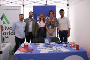 Cinco personas de pie detrás de una mesa con material promocional bajo una carpa. Las pancartas muestran logotipos.