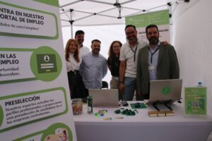 Un grupo de seis personas se encuentra bajo una carpa blanca en un evento de empleo. Están detrás de una mesa con computadoras portátiles, materiales promocionales y carteles informativos.