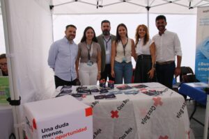 Seis personas están detrás de una mesa con material informativo y un mantel de la Cruz Roja bajo un dosel.