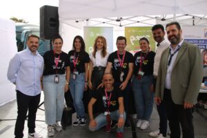 Un grupo de nueve personas de pie bajo una carpa blanca en un evento al aire libre. Algunos llevan camisetas negras con textos coloridos y otros llevan ropa informal o de negocios. Sonríen a la cámara.