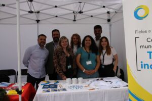 Un grupo de personas de pie bajo una carpa blanca sonríe a la cámara. En primer plano, una mesa muestra folletos y tazas.