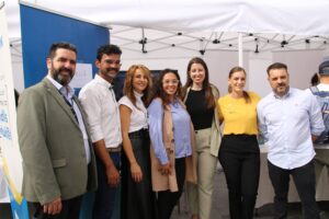 Siete personas están juntas, sonriendo, bajo un dosel blanco en un evento al aire libre. Algunas llevan ropa informal de negocios y hay una pancarta a la izquierda.