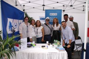 Un grupo de personas posando en un stand con una pancarta azul y materiales informativos bajo una carpa blanca.