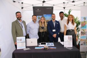 Un grupo de seis personas están de pie sonriendo detrás de una mesa con folletos y una computadora portátil, bajo un dosel.
