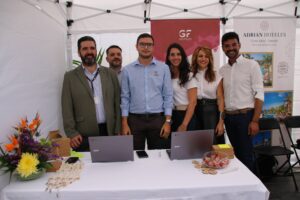 Un grupo de seis personas de pie detrás de una mesa con ordenadores portátiles, material promocional y flores bajo un dosel blanco. Al fondo se ven carteles de GF Hoteles y Adrian Hoteles.