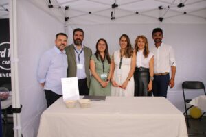Siete personas están de pie sonriendo dentro de una carpa blanca en un evento, con una mesa frente a ellos sosteniendo papeles y platos.