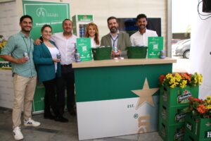 Un grupo de cinco personas sonrientes de pie detrás de un mostrador con latas y baldes de bebidas. El mostrador tiene una marca y un logotipo. Hay flores de colores y cajas de Heineken cerca.