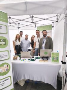 Personas de pie detrás de un stand con computadoras portátiles y materiales promocionales bajo un dosel.