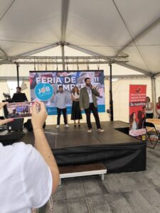 Tres personas en el escenario de una feria de empleo bajo una carpa. Un hombre sostiene un micrófono. Se ven a los asistentes en primer plano, incluido alguien que toma una foto con un teléfono inteligente.