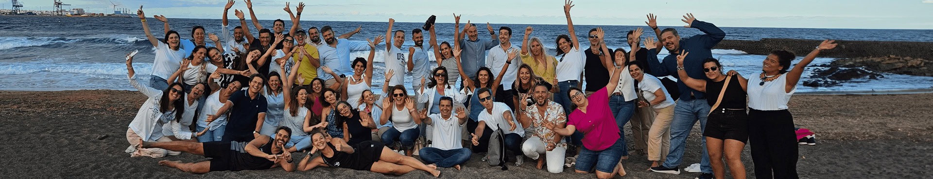 Un grupo diverso de personas posan juntas en una playa con el océano de fondo, muchas de ellas levantando los brazos y sonriendo para la foto durante el Encuentro anual de RRHH 2023.