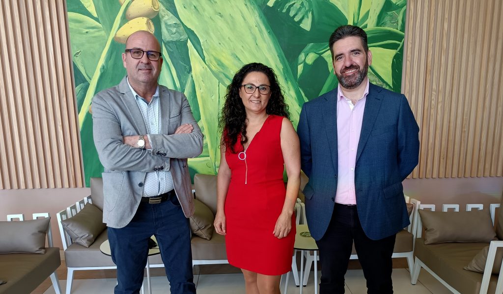 Tres personas posan para una fotografía en el interior. La mujer del centro, que representa al Grupo Macaronesian, lleva un vestido rojo y los dos hombres a cada lado llevan blazers. Están frente a un mural de hojas verdes.