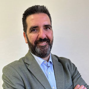 Un hombre con barba y bigote sonríe y está de pie con los brazos cruzados. Lleva una chaqueta gris claro sobre una camisa azul sobre un fondo liso. Para más información, contactar con gehocan.