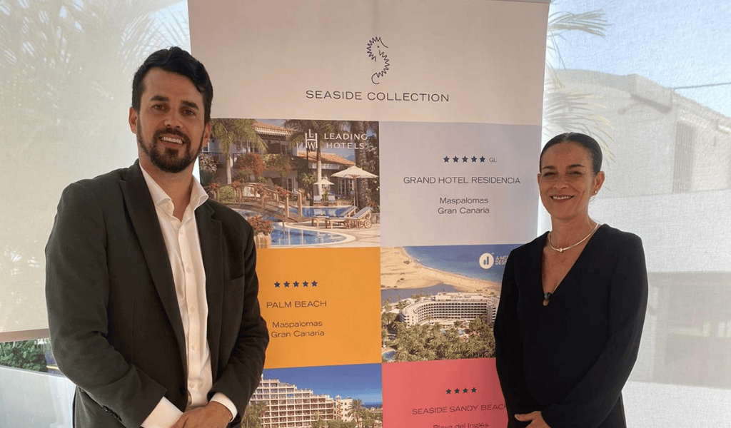 Dos personas se paran frente a un cartel de exhibición de los hoteles Seaside Collection. El banner, que muestra imágenes y nombres de los hoteles galardonados con el premio Gehocan 2022 en Gran Canaria, destaca la excelencia de esta estimada marca.