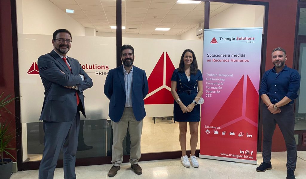 Cuatro personas se encuentran en una oficina con un letrero de RRHH de Triangle Solutions. Tres hombres vestidos de negocios y una mujer con un vestido azul forman el grupo. La pared de cristal detrás de ellos muestra los servicios de la empresa, incluido Gehocan, escritos en español.