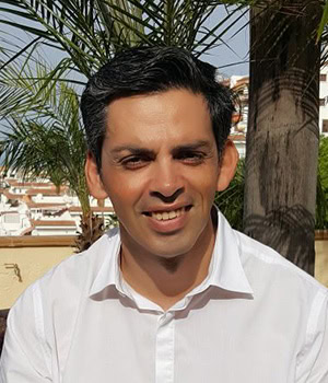 Un hombre de pelo corto y oscuro, vestido con una camisa blanca, sonríe al aire libre con palmeras y edificios al fondo. Para más detalles, contactar con gehocan.